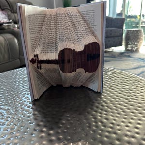 May include: A book folded into the shape of a violin. The book is open and the pages are folded to create the shape of the instrument. The book is sitting on a silver table.