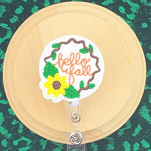 May include: A badge reel with a fall theme. The badge reel has a white background with the words "hello fall" in brown script, surrounded by a wreath of green leaves and a yellow sunflower. The reel is on a wooden surface.