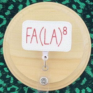Peut inclure: Un porte-badge avec le texte "FA(LA)8" brodé en rouge sur fond blanc. Le porte-badge est fixé à un cercle en bois, posé sur un tissu à imprimé léopard vert dans un cadre hexagonal en bois. Le mot "wordspapermagic" est en bas.