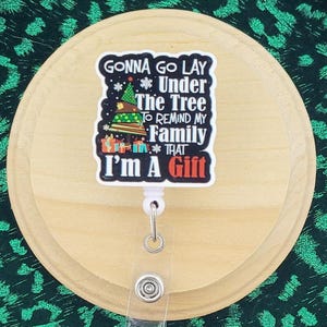 May include: A badge reel with a Christmas-themed design. The badge reel has a wooden backing and a green and black leopard print background. The badge reel has the text "Gonna go lay under the tree to remind my family that I'm a gift."