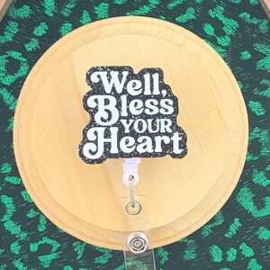 May include: A badge reel with the text "Well, Bless Your Heart" in a black and white design. The reel is attached to a wooden circle, set against a green leopard print background within a wooden hexagon frame. The words are in a decorative font.