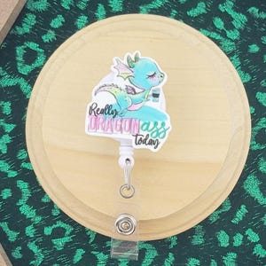 Peut inclure: Un porte-badge hexagonal en bois avec un fond à imprimé léopard vert. Un cercle en bois clair maintient un enrouleur de badge rétractable avec un dessin animé de dragon et le texte "Really Dragonass Today". Le bas du cadre porte le texte "wordspapermagic".