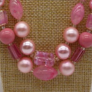 Puede incluir: Un collar de cuentas de color rosa de doble cadena con cuentas rosas grandes y pequeñas. El collar se muestra en un maniquí de tela marrón.