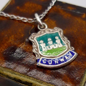 May include: A silver charm necklace with a blue, green, and white enamel pendant featuring the coat of arms of Conway, Wales. The charm is in a brown leather box.