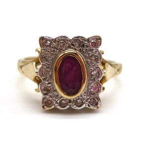 May include: A gold ring with an elaborate, rectangular setting. It features an oval ruby at the centre, encircled by small, clear stones. The ring's band is a plain gold design.