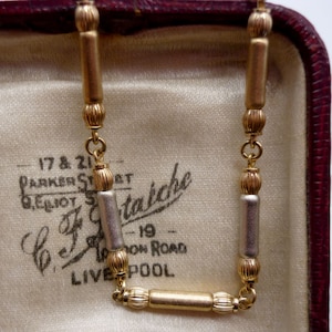 May include: A gold and silver chain necklace with a rectangular pendant. The chain is made of alternating gold and silver links. The pendant is a solid gold bar with a textured surface. The necklace is in a velvet-lined box with a label that reads "17 & 21 Parker Street, G. Elliot & Co. Statische, 19, London Road, Liverpool."