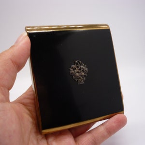 以下が含まれることがあります： 金色の縁取りと銀色の花の模様が施された黒い金属製のタバコケースです。