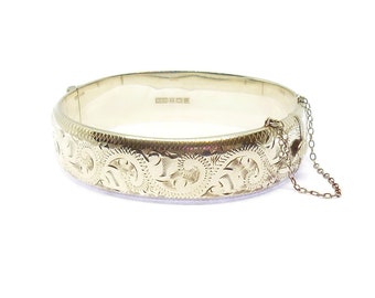 Könnte beinhalten: Ein goldenes Armreif-Armband mit einem gravierten Blumenmuster. Das Armband hat einen Scharnierverschluss und eine Sicherheitskette.