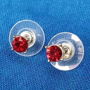 May include: A pair of stud earrings featuring bright red gemstones set in gold-coloured metal. The earrings are shown on clear, round backings against a blue backdrop.