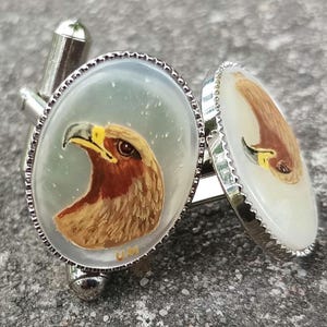 May include: A pair of silver cufflinks with a white background and a detailed painting of a golden eagle on one cufflink. The other cufflink has a white background with a partial view of the eagle's head.