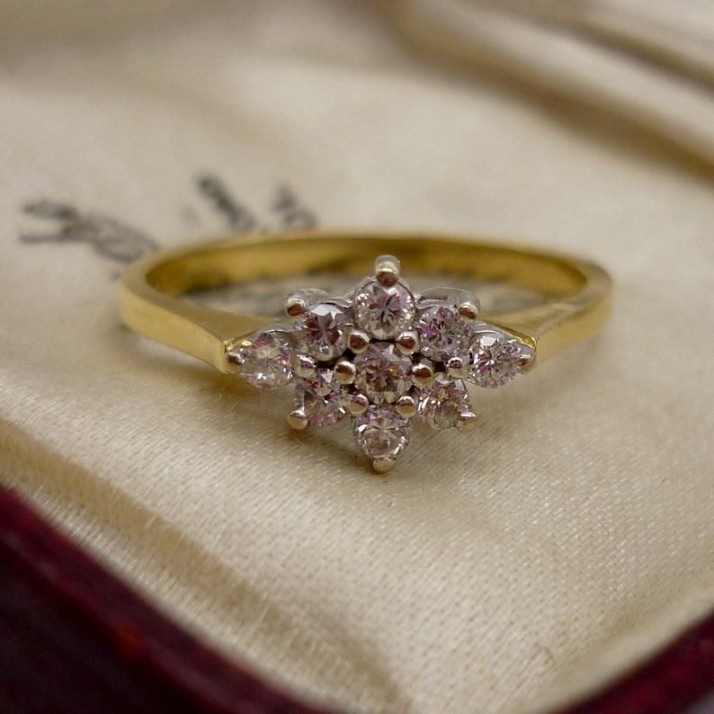 May include: A gold ring with a cluster of small, round diamonds forming a flower-like design. The ring is set on a light-colored fabric, showcasing the intricate details of the jewelry.