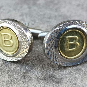May include: A pair of silver cufflinks with a gold-toned center featuring the letter "B".