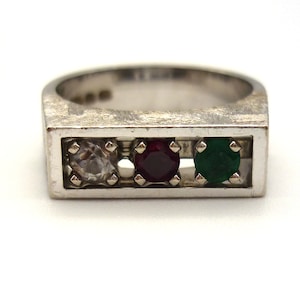 Puede incluir: Un anillo de plata con una banda rectangular y tres piedras preciosas. Las piedras preciosas son un diamante transparente, un rubí rojo y una esmeralda verde.