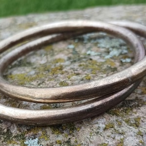 May include: Two bronze-coloured, circular bangles are intertwined, resting on a textured, moss-covered stone. The bangles have a slightly rough, metallic appearance, suggesting they are handmade or vintage. The background is blurred, with green grass visible.