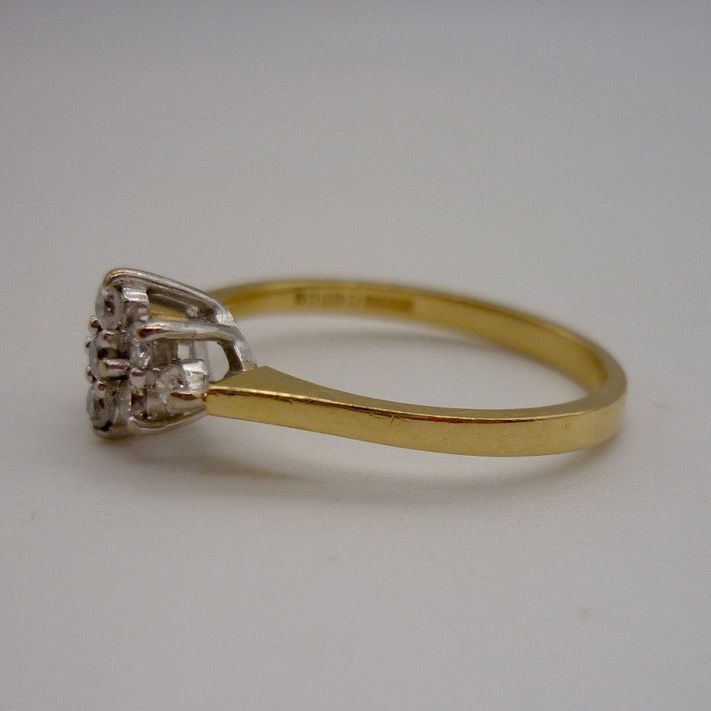 May include: A gold ring with a cluster of small, clear gemstones. The ring has a simple, elegant design with a gold band and a white metal setting for the gemstones. The gemstones are arranged in a floral or starburst pattern.