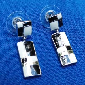 May include: A pair of geometric dangle earrings. Each earring has a square stud with black and white enamel squares, connected to a rectangular drop with a similar black and white enamel design. The earrings have a silver-tone metal finish.