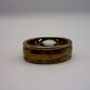 May include: A gold ring with a clear stone set in a decorative bezel. The ring has a wide band with a textured surface.