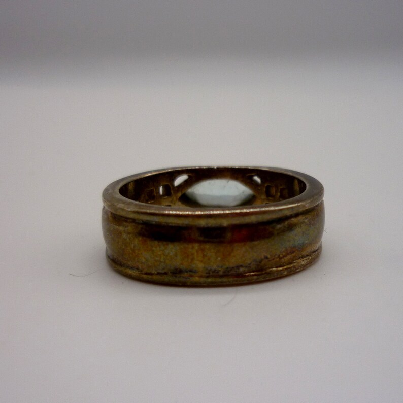 May include: A gold ring with a clear stone set in a decorative bezel. The ring has a wide band with a textured surface.