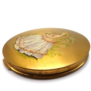 May include: A gold-toned compact mirror with a painted image of a woman in a white dress and a yellow hat. The woman is standing in a green field.