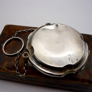 May include: A silver locket with a chain attached. The locket is round and has a smooth surface. It is sitting on a brown surface.
