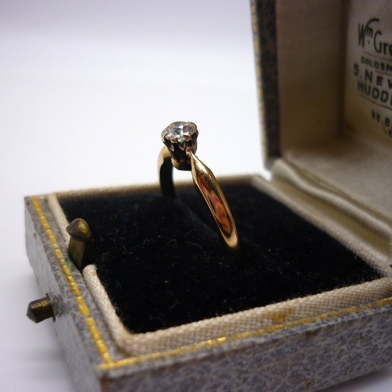 May include: A gold ring with a single diamond in a black velvet ring box. The box is lined with white satin and has a gold trim.