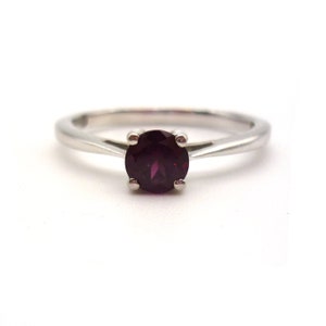 May include: A silver ring with a round, deep red gemstone. The gemstone is held in place by four prongs. The band is thin and smooth, with a simple, elegant design. The ring is set against a white background.