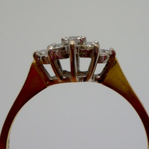 May include: Close-up of a gold ring with a cluster of three diamonds. The diamonds are set in a white metal, and the band is gold. The ring is set against a plain white background.