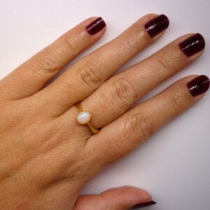 May include: A gold ring with a white opal gemstone. The ring is on a person's finger. The person's fingernails are painted a dark burgundy color.
