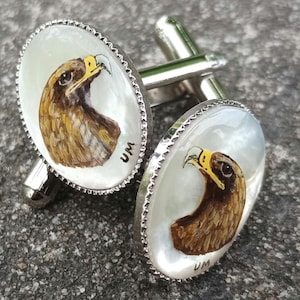 May include: A pair of silver cufflinks with a mother-of-pearl inlay featuring a detailed painting of a golden eagle. The cufflinks are oval shaped and have a silver rim.