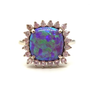 May include: A silver ring with a large, square-shaped opal gemstone surrounded by a halo of smaller, clear gemstones.