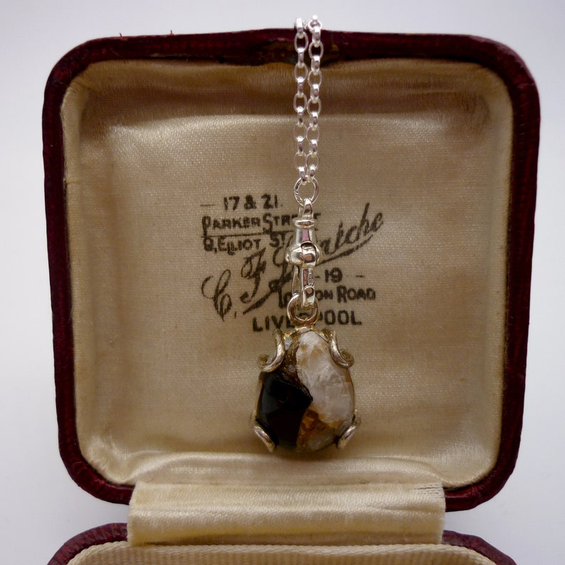 May include: A silver chain necklace with a pendant featuring a round, opaque, white and black stone set in a silver frame. The necklace is in a red velvet jewelry box with a white satin lining. The box has a label that reads "17 & 21. PARKER ST. BELLIOT ST. C.J.F. riche -19- PON ROAD LIVOOL."