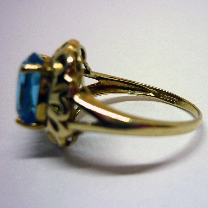May include: A gold ring with a large, oval-shaped blue gemstone set in a decorative, ornate setting. The ring is marked with the number "925" on the inside of the band.