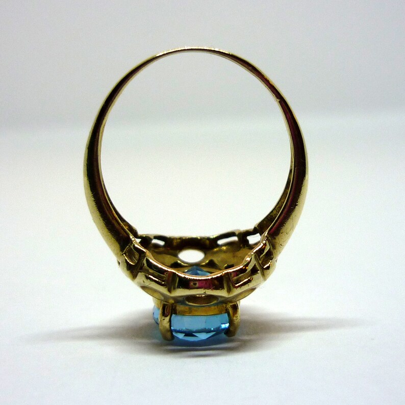 May include: A gold ring with a blue gemstone set in a delicate, ornate design. The ring is shown from the back, revealing the intricate details of the setting.
