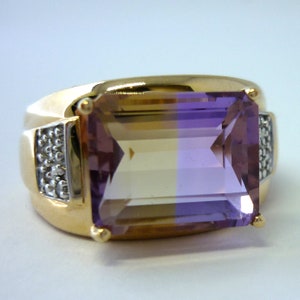 May include: A gold ring featuring a large, rectangular, bi-coloured gemstone. The gemstone is a light brown colour on the top half and a purple colour on the bottom half. The ring is set with small diamonds around the gemstone.