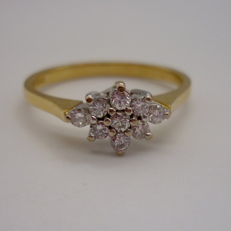 May include: A gold ring with a cluster of small, round diamonds forming a floral design. The diamonds are set in a white metal, contrasting with the yellow gold band. The ring is set against a white background.