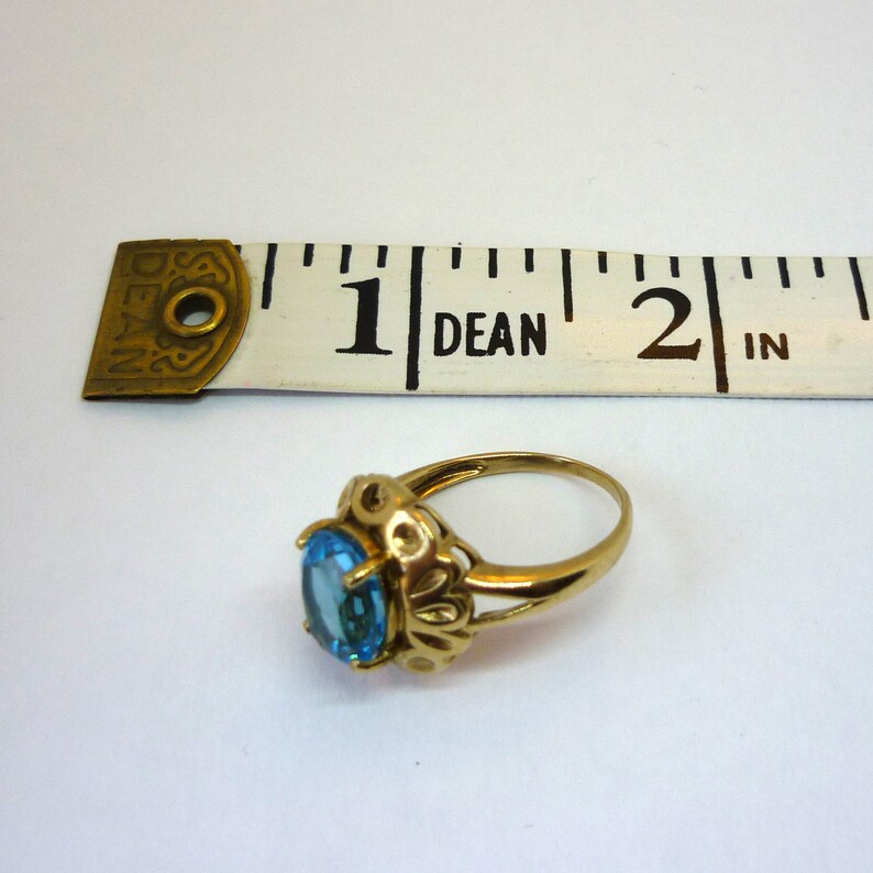 May include: A gold ring with a blue oval-shaped gemstone set in a decorative filigree design. The ring is measured against a ruler, showing it is slightly less than 1 inch in diameter.