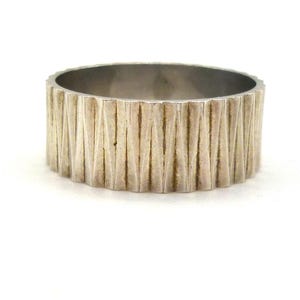 Puede incluir: Un anillo plateado con un diseño texturizado de ranuras verticales. El anillo tiene una banda ancha y un interior liso. El anillo se muestra sobre un fondo blanco, resaltando su brillo metálico y su detallada artesanía.