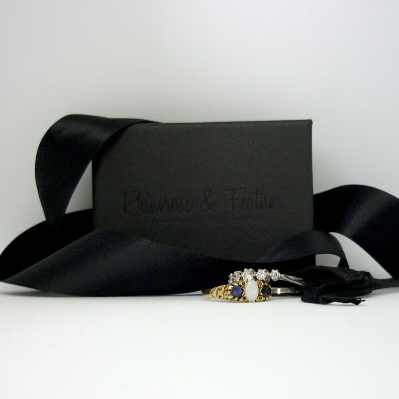 May include: A black jewelry box with a black satin ribbon and the text "Primrose & Feather" embossed on the lid. The box contains a gold ring with a white opal and blue sapphires.