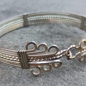 May include: Silver-toned bracelet with an intricate design. The bracelet features multiple parallel bands, with decorative elements including twisted wire and ornate clasps. The clasp has a series of loops and swirls. The jewelry is displayed on a gray surface.