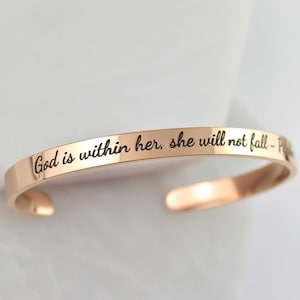 Puede incluir: Una pulsera tipo brazalete de metal en tono oro rosa con acabado pulido. La superficie exterior presenta el texto grabado en negro "God is within her, she will not fall - Psalm 46:5" en letra cursiva. La pulsera tiene un diseño abierto.