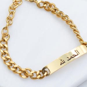 May include: A gold-toned chain link bracelet featuring a central rectangular metal plate engraved with the Arabic script text Alhamdullilah. The bracelet includes a lobster claw clasp closure and a series of interlocking oval links in a polished finish.
