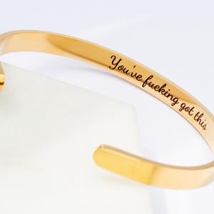 Könnte beinhalten: Offener Armreif in Goldfarbe. Der Armreif trägt den eingravierten Text "You've f*cking got this". Der Armreif liegt auf einer weißen Oberfläche. Der Armreif ist ein schlichtes, elegantes Design.
