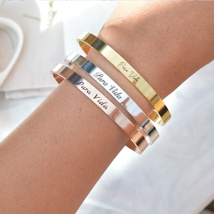 May include: Three gold, silver and rose gold bangle bracelets with the phrase "Pura Vida" engraved on each. The bracelets are stacked on a person's wrist.
