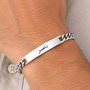 May include: A silver-toned metal chain bracelet featuring a flat, rectangular identification plate in the center. The plate is engraved with the Arabic name Nasser in black lettering. The bracelet consists of interlocking curb chain links.