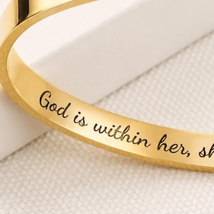 Può includere: Un bracciale rigido color oro dal design aperto con un piccolo intaglio a forma di cuore su un'estremità. La superficie interna è incisa con il testo nero, God is within her, she will not fall Psalm 46:5, in un carattere corsivo.