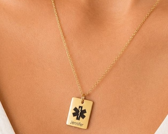 May include: A gold necklace with a rectangular pendant. The pendant displays a black medical symbol and the name "Jennifer" engraved beneath. The necklace is worn against a white top.