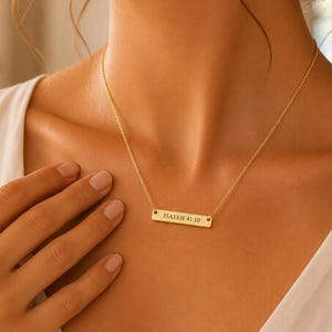 May include: A gold-toned bar necklace featuring a delicate chain and a rectangular pendant engraved with the text Isaiah 41:10. The piece rests against the collarbone, showcasing a polished metallic finish and a simple, clean design.