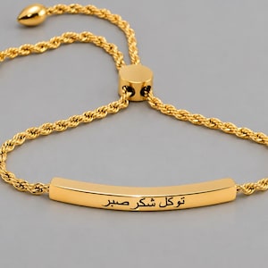Könnte beinhalten: Ein verstellbares goldfarbenes Armband mit einer gebogenen Metallstange, die mit dem arabischen Text "توكل شكر صبر" graviert ist. Das Armband besteht aus einer gedrehten Kette in Seiloptik und verfügt über einen Schiebeverschluss zur Größenanpassung.