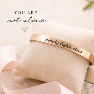 Op de afbeelding: Een roségouden armband met de gegraveerde tekst "nobody fights alone" rustend op een beige fluwelen kussen. Daarnaast ligt een kaartje met een harticoon en de gedrukte woorden "YOU ARE not alone." en "STRENGTH. COURAGE. HOPE."