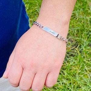 May include: A silver-toned metal chain link bracelet featuring a flat, rectangular identification plate in the center. The plate is engraved with the Arabic name "ناصر" in black lettering. The bracelet has a polished finish and a secure clasp.